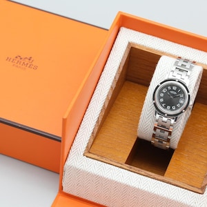 May include: A silver-tone wristwatch with a black circular dial featuring white Arabic numerals and a date window, presented on a metallic link bracelet with H-shaped links, housed in an orange box with the text HERMES PARIS printed on the lid.