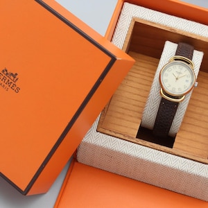 May include: A gold-tone wristwatch with a round cream-colored dial, Arabic numerals, and a dark brown textured leather strap. The watch is presented inside a square orange box with the Hermes Paris logo and a light-colored herringbone fabric interior.