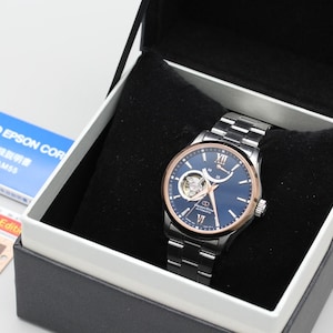May include: A stainless steel wristwatch with a blue face and gold-tone accents, presented in a black box with a white interior. The watch features a silver band and is accompanied by a Seiko Epson Corp. booklet.