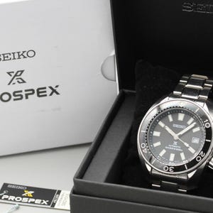 以下が含まれることがあります： 黒い文字盤とシルバーのステンレススチールバンドを備えたセイコープロスペックス自動巻き腕時計。黒い箱に入っています。時計には回転ベゼルがあり、黒いクッションの上に表示されています。セイコーとプロスペックスのロゴが見えます。