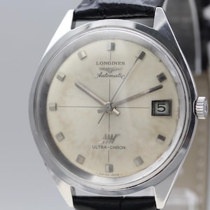 Op de afbeelding: Een vintage Longines Ultra-Chron automatisch polshorloge. Het horloge heeft een zilveren wijzerplaat met rechthoekige uurmarkeringen, een datumvenster op 3 uur en zilveren wijzers. De kast is van roestvrij staal en de band van zwart leer.
