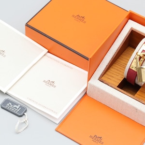 May include: An open orange box with a luxury watch inside. The watch has a red leather strap and a gold-colored padlock clasp. The box also contains white booklets and a tag with the brand's logo.