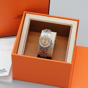 May include: A silver-tone wristwatch with a round beige dial and Arabic numerals, featuring a metal link bracelet. The watch is presented inside an open orange box with a wooden interior, accompanied by a white instruction booklet with Hermes Paris branding.