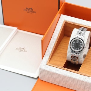 May include: A silver-tone wristwatch with a black circular dial featuring white Arabic numerals and the Hermes Paris logo. The watch has a metallic link bracelet and is presented in an orange box with a white herringbone interior and booklets.