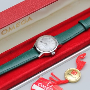 May include: A vintage Omega wristwatch with a silver dial and a green leather strap. The watch is presented in a red box with the brand name "OMEGA" printed in gold. A gold-colored tag with the Omega logo is attached.