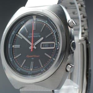 Puede incluir: Un reloj Seiko 5 Sports Speed-Timer vintage con caja y correa de acero inoxidable. El reloj tiene una esfera negra con detalles rojos y blancos, una pantalla de día y fecha y un logotipo Speed-Timer. La esfera mide aproximadamente 3,8 cm de diámetro.