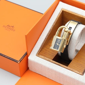 May include: A gold-tone padlock-shaped wristwatch with a black dial and a black leather strap, presented in an orange box with the text HERMES PARIS. The watch features a gold-tone lock mechanism and is displayed on a white fabric cushion.