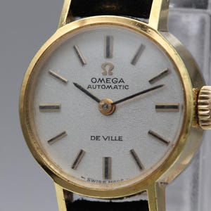 May include: A gold-toned Omega De Ville automatic wristwatch with a white dial. The watch features black hands and hour markers, and the words "OMEGA AUTOMATIC" and "DE VILLE" are printed on the dial. The watch has a black leather strap.
