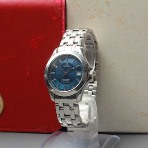 May include: A stainless steel Omega Seamaster watch with a blue dial. The watch has a date window and a silver link bracelet. The watch is displayed on a clear stand, with a red leather case and a box in the background.