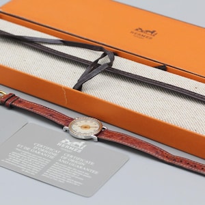 May include: A wristwatch with a round silver-tone case, a white dial with gold-tone numerals, and a textured reddish-brown leather strap. The watch is accompanied by an orange presentation box, a dark brown leather strap, and a certificate of origin.