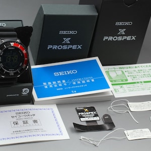 May include: A black Seiko Prospex digital watch with a red and black bezel, presented in its original box with paperwork. The watch face displays the time. Includes instruction manuals and tags.