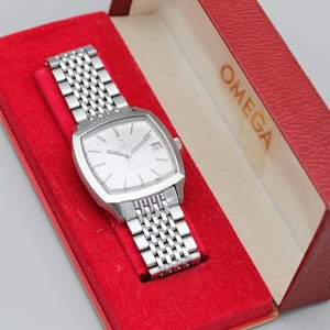 May include: A silver-toned Omega wristwatch with a square face and a linked band, presented in a red box with the brand name printed in gold. The watch is set against a red velvet interior.