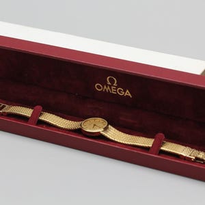 May include: A gold-tone Omega wristwatch with a round face and a textured mesh link bracelet, presented inside a rectangular burgundy velvet-lined display case with the Omega logo printed in gold on the interior lid, accompanied by a white outer box.