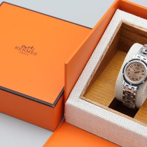 May include: A silver-tone wristwatch with a round face and metallic link bracelet, displayed on a white fabric cushion inside a wooden insert. The watch is housed in an open orange box with the text HERMES PARIS printed on the lid.