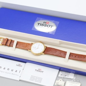 May include: A Tissot analog quartz watch with a gold-tone round case, white dial, and brown textured leather strap. The watch is presented in a blue and white rectangular box with a Tissot logo, accompanied by a manual and small white tags.