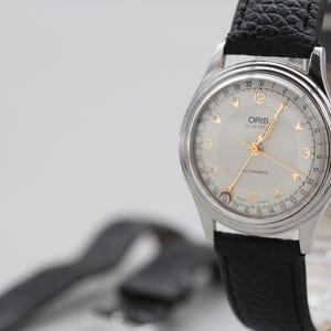 Może przedstawiać: Srebrny automatyczny zegarek Oris z jasnoszarą tarczą i złotymi wskazówkami. Zegarek ma czarny skórzany pasek i jest prezentowany na białym tle. Na tarczy zegarka widnieje napis "ORIS" i "17 JEWELS".