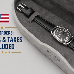 May include: A wristwatch with a black leather strap and a silver-toned rectangular case featuring a black dial with white Arabic numerals. The watch is presented inside a grey felt-lined storage case with the brand name Hermes Horloger embossed.
