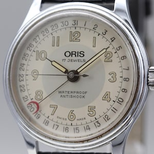 Könnte beinhalten: Eine silberfarbene Oris Armbanduhr mit cremefarbenem Zifferblatt, arabischen Ziffern, einem Datumszeiger mit roter Mondsichelspitze und dem Text Oris 17 Jewels Waterproof Antishock Swiss Made. Die Uhr hat ein Gehäuse aus poliertem Metall.