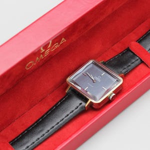 May include: A vintage Omega De Ville watch with a square face and black leather band, presented in a red box. The watch has a gold-toned case and a dark gray dial. The box is red with the Omega logo.