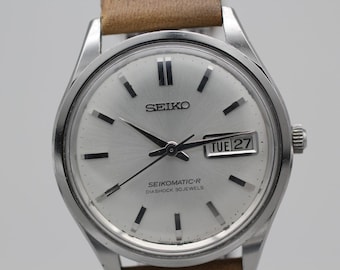 May include: A silver Seiko watch with a light brown leather band. The watch face is silver with black markers and hands. The date reads "TUE 27". The watch is a Seiko Seikomatic-R Diashock 30 Jewels.
