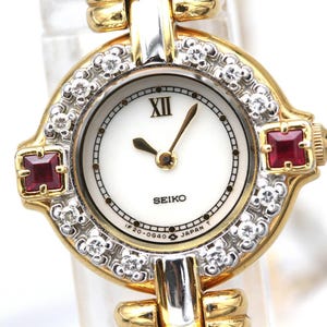 May include: A Seiko wristwatch featuring a round white dial with a Roman numeral XII at the top, gold hands, and a bezel set with clear stones and two square red stones. The watch case is gold-toned with silver accents and a link bracelet.