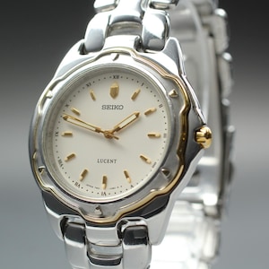 Puede incluir: Un reloj Seiko Lucent plateado con esfera blanca y agujas y detalles dorados. El reloj presenta una correa de acero inoxidable y una esfera redonda con números romanos.