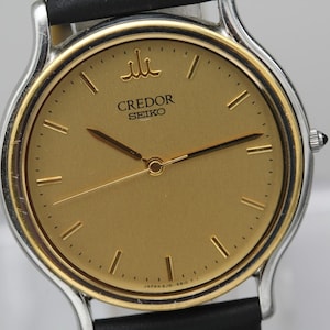 May include: A Credor Seiko wristwatch featuring a round gold-toned dial with thin gold hour markers and matching hands. The watch has a silver-toned bezel and a black leather strap. The Credor logo is positioned at the twelve o'clock mark.