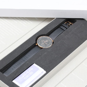 May include: A black wristwatch with a rose gold-colored bezel and a black leather band is presented in a gray box. The watch face has a dark background with multiple sub-dials. The watch is packaged in a white box.