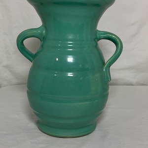 Puede incluir: Un jarrón de cerámica de color turquesa con dos asas en forma de lazo. El jarrón tiene un borde ancho y acampanado y un cuerpo redondeado con estrías horizontales. El acabado brillante refleja la luz.