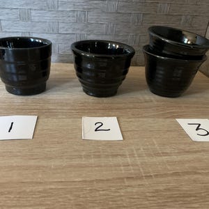 Könnte beinhalten: Drei glänzend schwarze Keramikschalen mit geripptem, konischem Design. Die Schalen sind in einer Reihe angeordnet, wobei die ersten beiden einzeln stehen und die dritte Schale auf einer anderen gestapelt ist. Jede Schale ist mit einer kleinen weißen Karte mit der Nummer 1, 2 oder 3 gekennzeichnet.