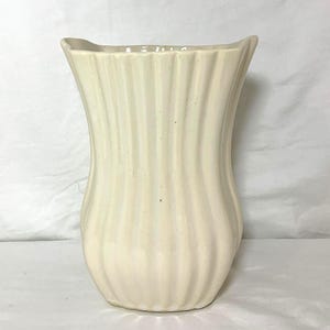 Puede incluir: Jarrón de cerámica color crema con diseño acanalado. El jarrón tiene una abertura ancha, ligeramente curvada y un cuerpo suavemente curvado. El acanalado vertical crea una superficie texturizada. Una pieza decorativa para el hogar.