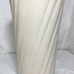 Puede incluir: Un jarrón alto de cerámica blanquecina con un diseño acanalado en espiral. El jarrón tiene una parte superior ligeramente ensanchada y una base lisa y redondeada. El color neutro y la superficie texturizada lo convierten en un artículo decorativo versátil.