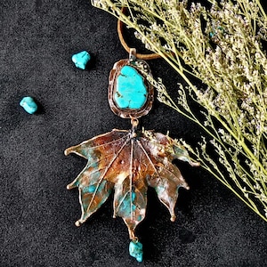 May include: A handcrafted necklace featuring a large, copper-toned leaf pendant with turquoise accents. The necklace includes a turquoise stone and a turquoise stone pendant. The necklace is on a brown cord, with a black background.