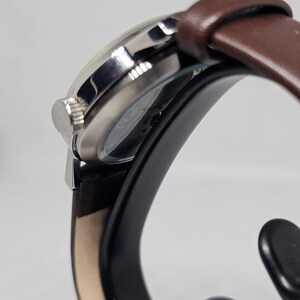 K&ouml;nnte beinhalten: Eine silberfarbene Armbanduhr mit braunem Lederarmband. Das Zifferblatt der Uhr ist sichtbar, und die Krone befindet sich an der Seite. Die Uhr wird auf einem schwarzen St&auml;nder vor wei&szlig;em Hintergrund pr&auml;sentiert. Das Armband ist etwa 2 cm breit.
