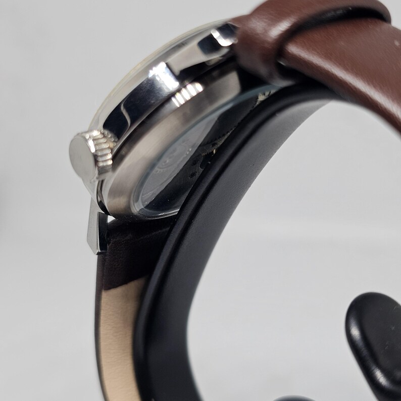 K&ouml;nnte beinhalten: Eine silberfarbene Armbanduhr mit braunem Lederarmband. Das Zifferblatt der Uhr ist sichtbar, und die Krone befindet sich an der Seite. Die Uhr wird auf einem schwarzen St&auml;nder vor wei&szlig;em Hintergrund pr&auml;sentiert. Das Armband ist etwa 2 cm breit.
