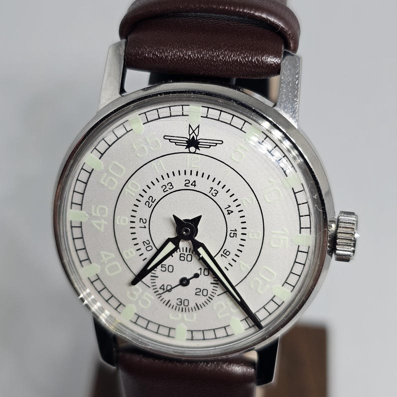 K&ouml;nnte beinhalten: Eine silberfarbene Armbanduhr mit wei&szlig;em Zifferblatt, schwarzen Zeigern und einem braunen Lederarmband. Das Zifferblatt zeigt eine 24-Stunden-Skala, eine Sekundenanzeige und ein Vogellogo. Die Uhr hat eine Krone auf der rechten Seite.