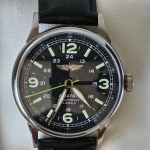 Può includere: Un orologio da polso con quadrante nero, cassa argentata e cinturino in pelle nera. Il quadrante presenta numeri bianchi, una lancetta dei secondi con punta verde e la scritta "MATON YOUR SCALE 19 JEWELS". L'orologio ha un design classico in stile pilota.