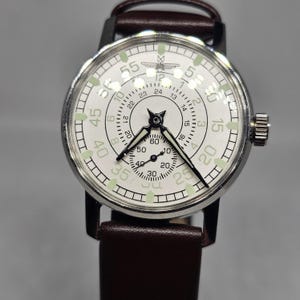 K&ouml;nnte beinhalten: Eine Vintage-Armbanduhr mit wei&szlig;em Zifferblatt, schwarzen Zeigern und einem braunen Lederarmband. Die Uhr hat ein silberfarbenes Geh&auml;use und ein zweites Zifferblatt. Das Zifferblatt zeigt Zahlen und Markierungen in Schwarz und Gr&uuml;n.