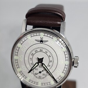 K&ouml;nnte beinhalten: Silberfarbene Armbanduhr mit braunem Lederarmband. Das Zifferblatt der Uhr hat schwarze Zeiger, ein zweites Zifferblatt und ein Design oben. Die Uhr hat eine silberne L&uuml;nette und eine Krone an der Seite.