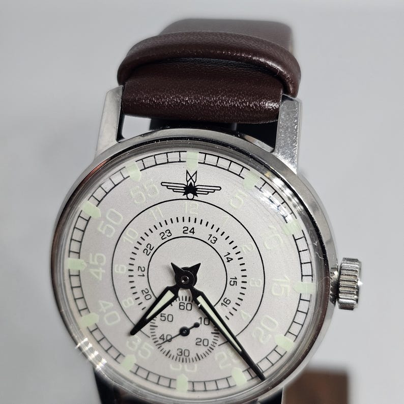 K&ouml;nnte beinhalten: Silberfarbene Armbanduhr mit braunem Lederarmband. Das Zifferblatt der Uhr hat schwarze Zeiger, ein zweites Zifferblatt und ein Design oben. Die Uhr hat eine silberne L&uuml;nette und eine Krone an der Seite.