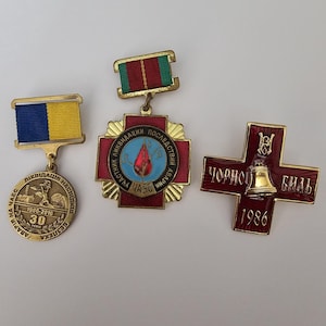 Könnte beinhalten: Drei Vintage-Medaillen. Die erste hat ein blau-gelbes Band und einen runden Anhänger mit Text. Die zweite ist eine rot-goldene kreuzförmige Medaille mit Text. Die dritte ist eine rot-goldene kreuzförmige Medaille mit einer Glocke und Text.
