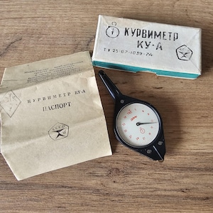 Puede incluir: Un instrumento Kurvímetro KU-A vintage en blanco y negro con una esfera circular, un mango negro y un documento en papel y una caja con texto cirílico. La esfera tiene números y un indicador rojo.
