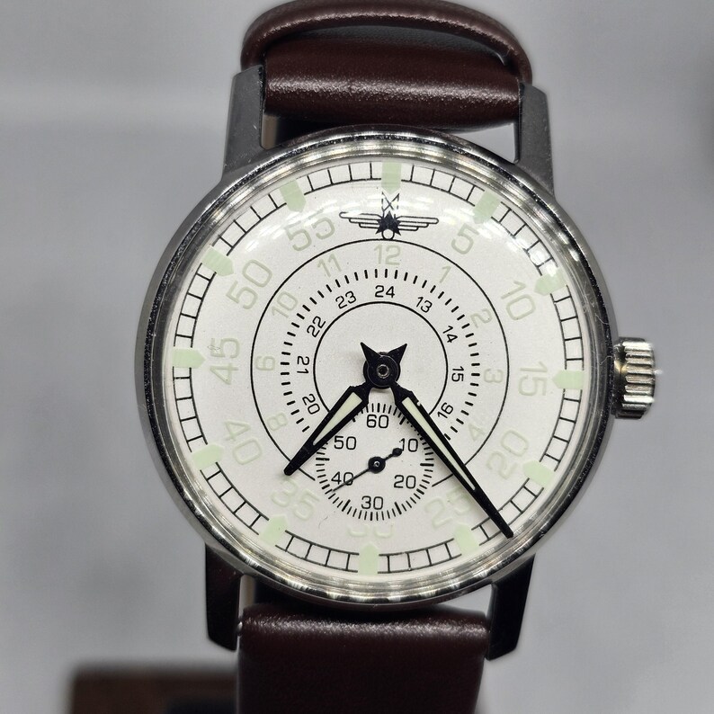 K&ouml;nnte beinhalten: Eine Vintage-Armbanduhr mit wei&szlig;em Zifferblatt, schwarzen Zeigern und einem braunen Lederarmband. Die Uhr hat ein silberfarbenes Geh&auml;use und verf&uuml;gt &uuml;ber mehrere Zifferbl&auml;tter und Markierungen zur Zeitmessung. Das Zifferblatt enth&auml;lt die Zahlen 5, 10, 15, 20, 30, 40, 50 und 55.