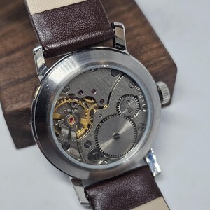 K&ouml;nnte beinhalten: Nahaufnahme einer Vintage-Armbanduhr mit silbernem Geh&auml;use und braunem Lederarmband. Das komplizierte mechanische Uhrwerk der Uhr mit goldenen Zahnr&auml;dern und Rubinjuwelen ist durch den transparenten Boden sichtbar. Das Armband ist 20 mm breit.