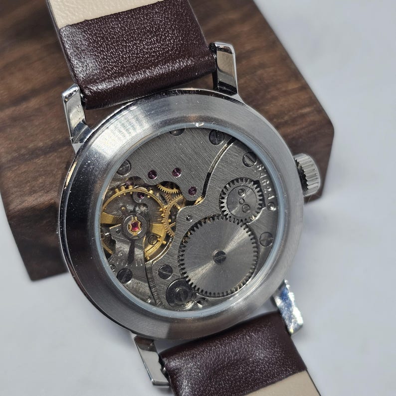 K&ouml;nnte beinhalten: Nahaufnahme einer Vintage-Armbanduhr mit silbernem Geh&auml;use und braunem Lederarmband. Das komplizierte mechanische Uhrwerk der Uhr mit goldenen Zahnr&auml;dern und Rubinjuwelen ist durch den transparenten Boden sichtbar. Das Armband ist 20 mm breit.