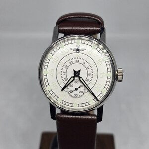 K&ouml;nnte beinhalten: Eine Vintage-Armbanduhr mit wei&szlig;em Zifferblatt, schwarzen Zeigern und einem braunen Lederarmband. Das Zifferblatt zeigt schwarze Ziffern und Markierungen mit einem kleinen Emblem oben. Die Uhr hat ein silberfarbenes Geh&auml;use und eine Krone an der Seite.