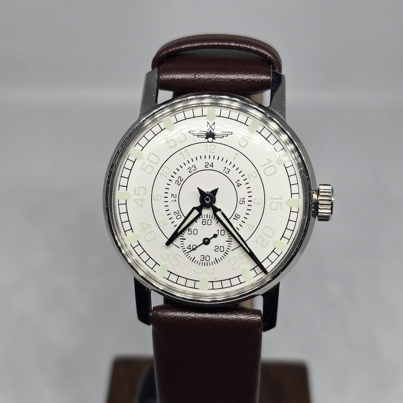 K&ouml;nnte beinhalten: Eine Vintage-Armbanduhr mit wei&szlig;em Zifferblatt, schwarzen Zeigern und einem braunen Lederarmband. Das Zifferblatt zeigt schwarze Ziffern und Markierungen mit einem kleinen Emblem oben. Die Uhr hat ein silberfarbenes Geh&auml;use und eine Krone an der Seite.