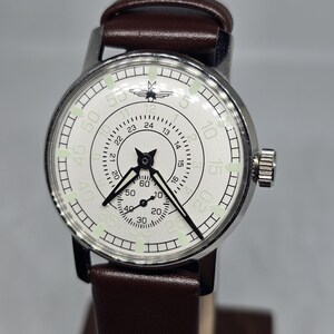 K&ouml;nnte beinhalten: Eine Vintage-Armbanduhr mit wei&szlig;em Zifferblatt, schwarzen Zeigern und einem braunen Lederarmband. Die Uhr hat ein silberfarbenes Geh&auml;use und ein kleines Hilfszifferblatt. Das Zifferblatt zeigt Zahlen und Markierungen zur Zeitmessung.