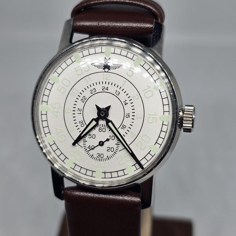 K&ouml;nnte beinhalten: Eine Vintage-Armbanduhr mit wei&szlig;em Zifferblatt, schwarzen Zeigern und einem braunen Lederarmband. Die Uhr hat ein silberfarbenes Geh&auml;use und ein kleines Hilfszifferblatt. Das Zifferblatt zeigt Zahlen und Markierungen zur Zeitmessung.