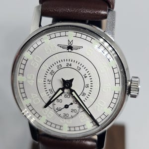 K&ouml;nnte beinhalten: Eine silberfarbene Armbanduhr mit einem braunen Lederarmband. Das Zifferblatt der Uhr ist wei&szlig; mit schwarzen Ziffern und Zeigern sowie einem kleinen Sekundenzeiger. Die Uhr hat eine Krone zum Aufziehen und Einstellen der Zeit. Das Armband ist ca. 20 cm lang.